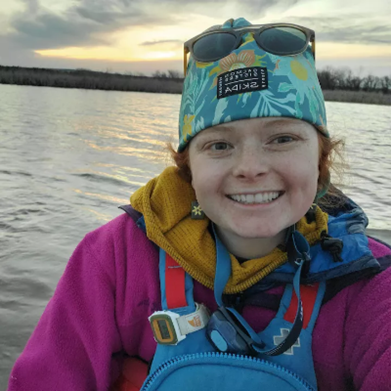 The image shows a woman smiling at the camera while on a body of water. She is wearing a hat, sunglasses, and a pink jacket. The background shows a sunset over the water. The woman appears to be enjoying herself.